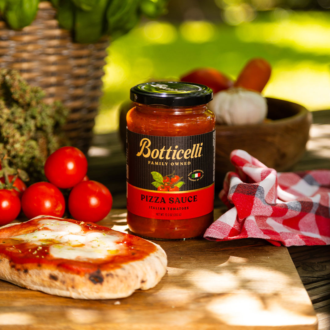 Botticelli pizza sauce jar with a slice of bread spread with sauce and tomatoes on a wooden table.